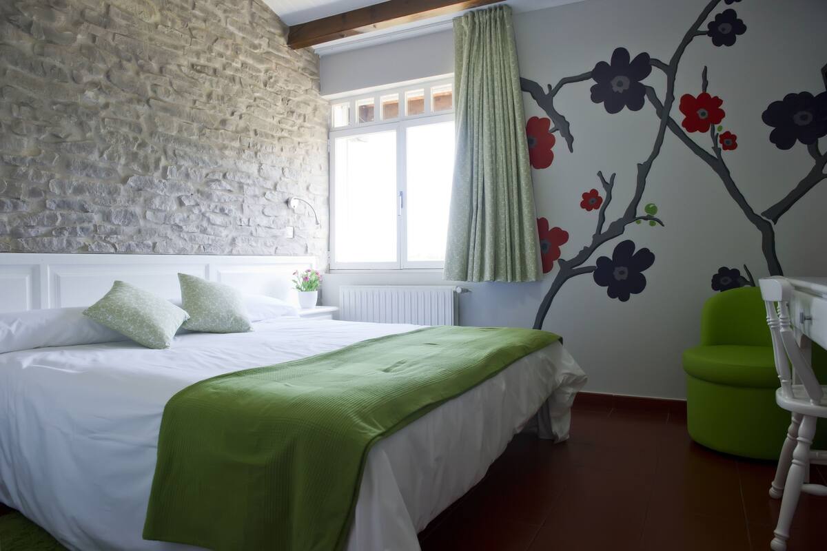 Un dormitorio con pared de piedra, cama con ropa de cama blanca y detalles verdes, una ventana con cortina verde y un mural floral.