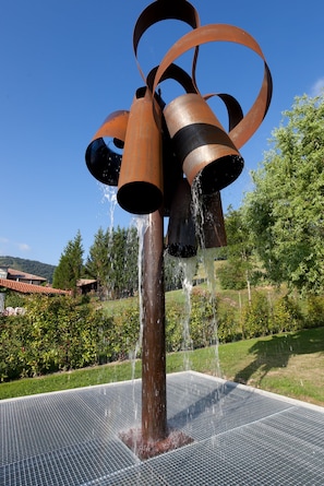 Una escultura de metal con agua que fluye desde la parte superior, ubicada en un área ajardinada con árboles y cielo despejado.
