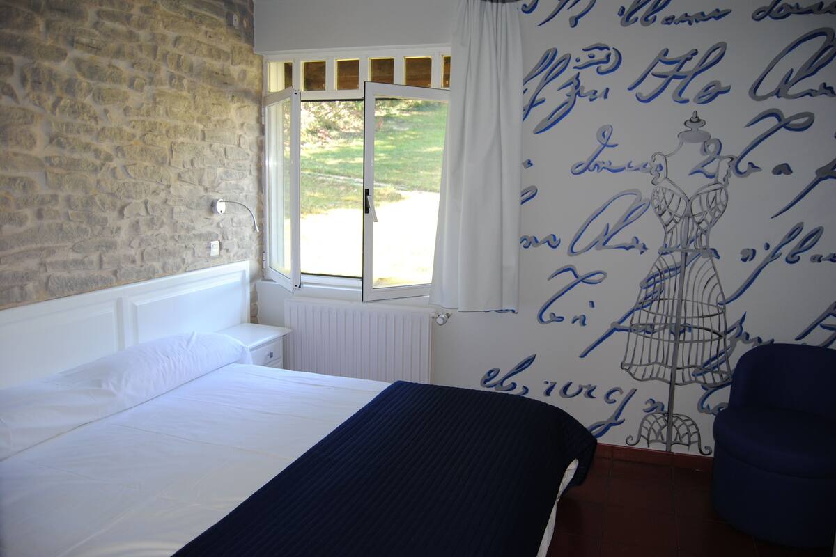 Un dormitorio con pared de piedra, una ventana con cortinas blancas, una cama con ropa de cama blanca y una silla azul.
