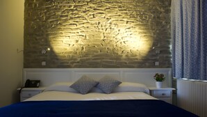 A bed with a stone wall backdrop, a bedside table with a phone, and a nightstand with a plant. - Izalde (Ayala)