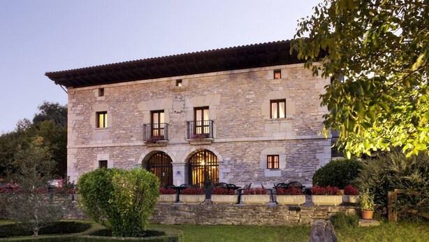 A stone building with a tiled roof, arched windows, and a well-maintained garden with hedges and potted plants. - Izalde (Ayala)
