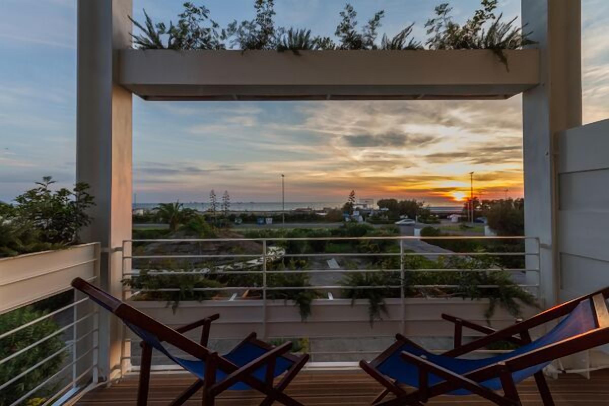 A balcony with two blue lounge chairs, a view of the sunset, and greenery.