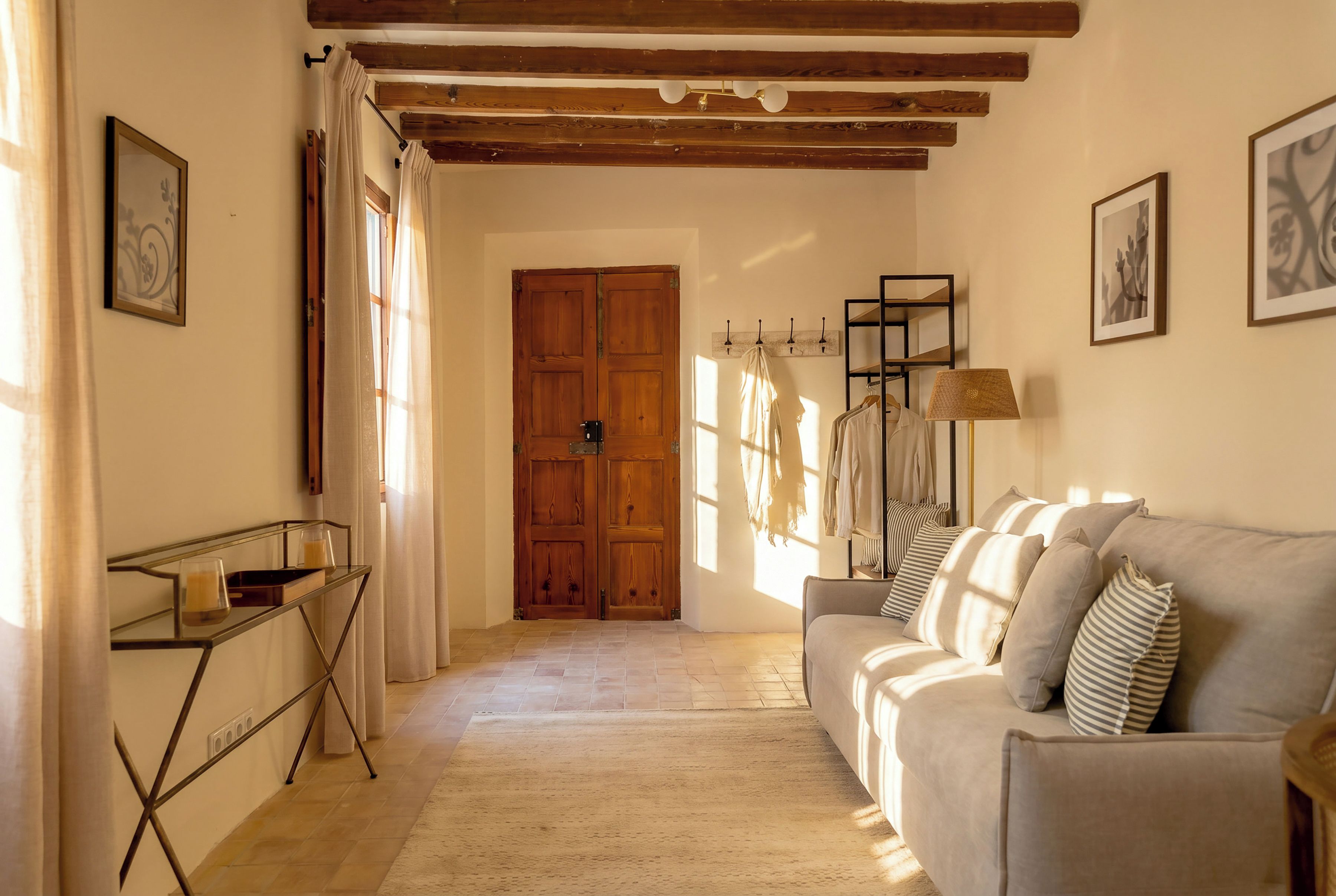 A hallway with a wooden door, a glass table with a candle, a sofa with striped pillows, and a wall-mounted lamp.