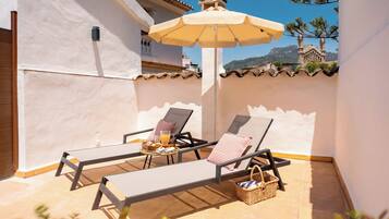 A sunlit patio with two lounge chairs, a table with a tray of food, and a large umbrella.
