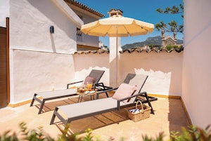 Eine sonnendurchflutete Terrasse mit zwei Liegestühlen, einem Tisch und einer Speiseplatte unter einem großen Sonnenschirm.