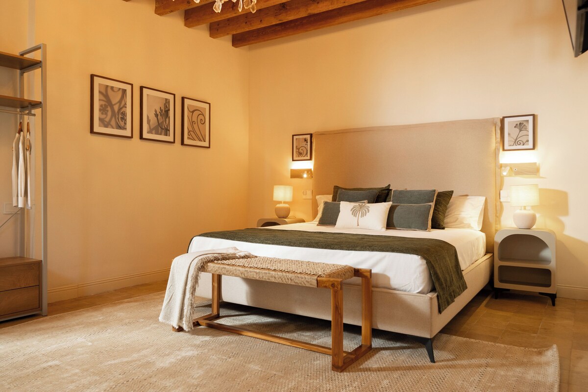A bedroom with a bed, bedside lamps, a bench, and framed artwork on the wall.