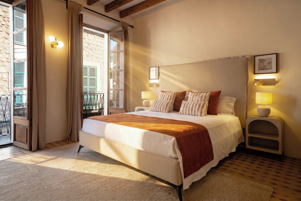 A bedroom with a bed, bedside lamps, a window with shutters, and a view of the outside.