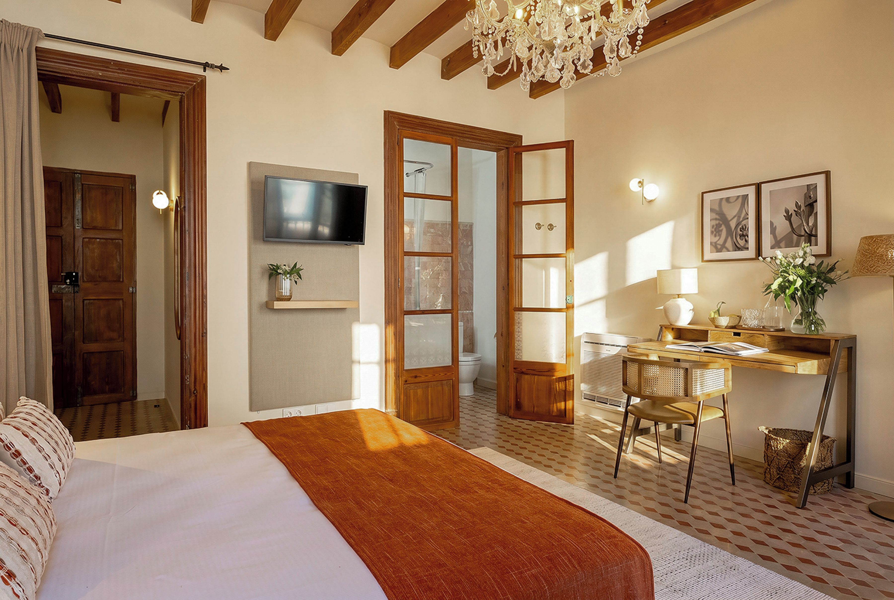 A bedroom with a bed, a desk, a chair, a television, and a chandelier.