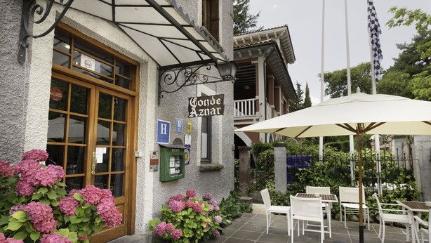 A building with a sign reading "Conde Aznar," outdoor seating with white chairs and tables, and a large umbrella. - Conde Aznar (Jaca)