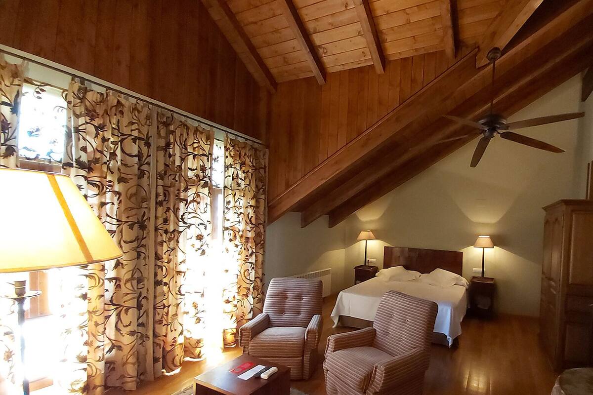 A cozy room with wooden beams, a ceiling fan, two bedside lamps, a bed, and a chair.