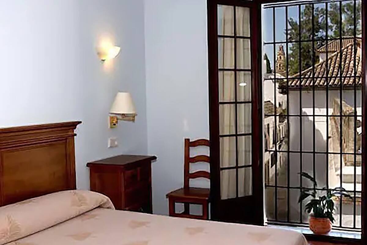 A bedroom with a bed, a nightstand, a chair, a lamp, and a view through a door to the outside.