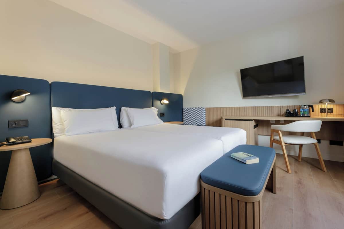A modern hotel room with a large bed, bedside tables, a desk with a chair, a flat-screen TV, and a small reading area.