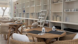 Un restaurante con mesas de madera redondas y sillas de mimbre, estanterías blancas con cristalería y vista al mar.