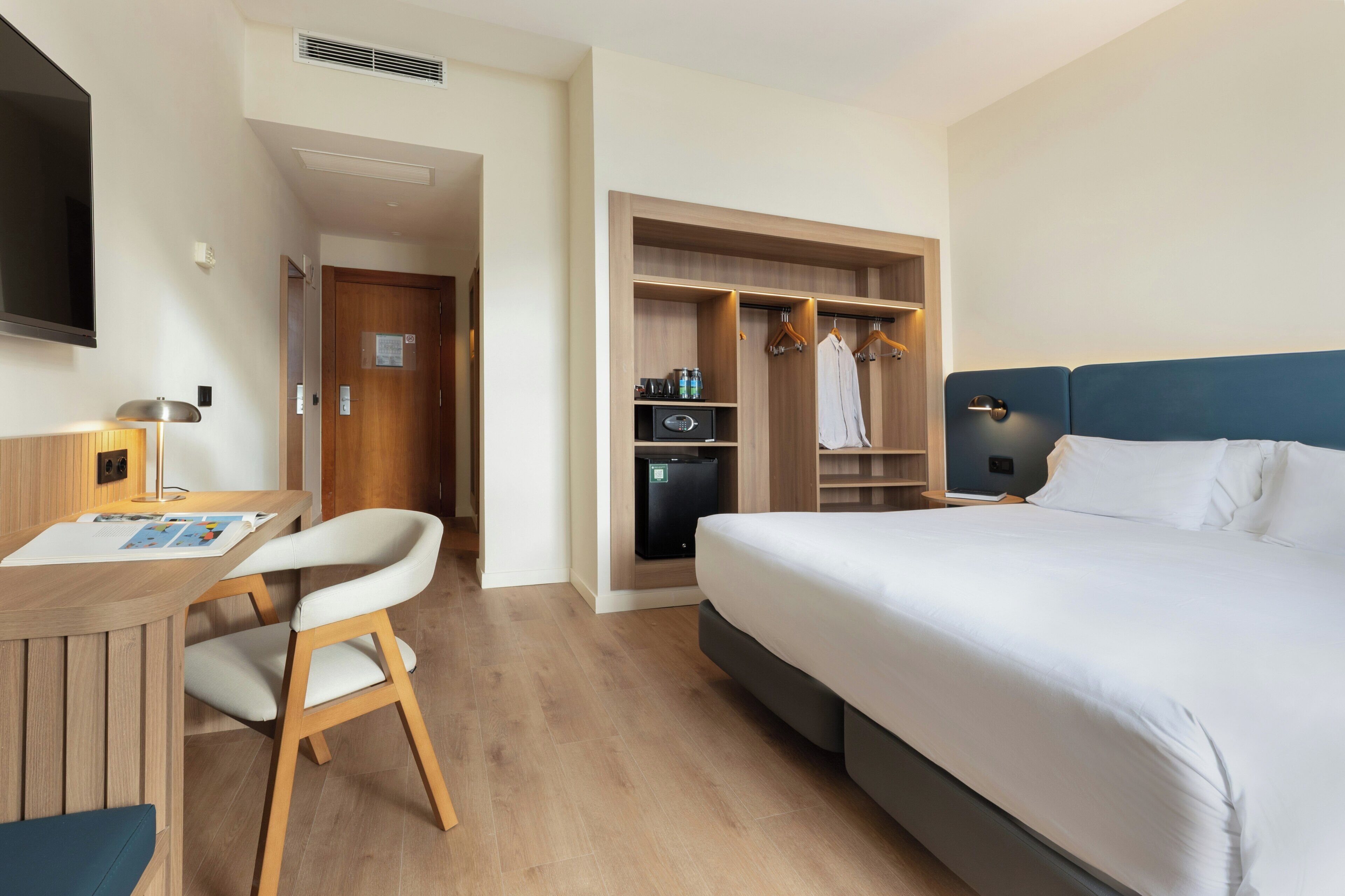 A hotel room with a bed, desk, chair, wardrobe, and a TV.