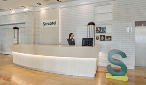 A reception desk with a sign reading "Sercotel," a woman standing behind it, and a large decorative letter 'S' on the floor. - Sercotel Kalma Sitges (Sitges)
