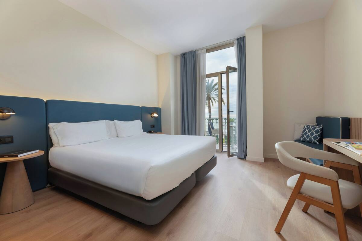 A hotel room with a large bed, a desk, and a chair. There is a balcony with a view of palm trees.