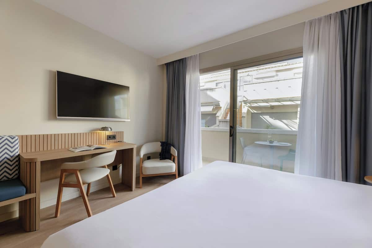 A hotel room with a large bed, a desk with a chair, a TV mounted on the wall, and a balcony with curtains.