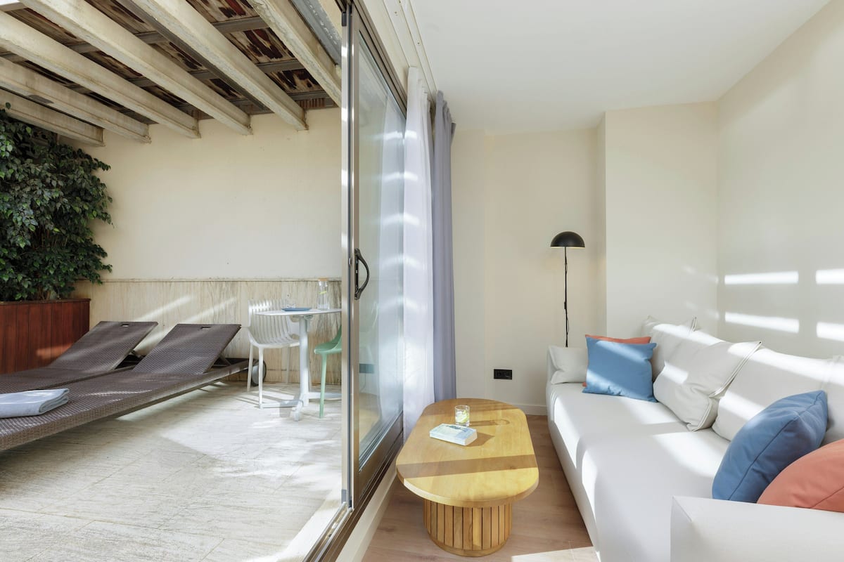 A modern room with a glass door leading to a balcony, a white sofa with colorful cushions, a wooden coffee table, and a green chair.