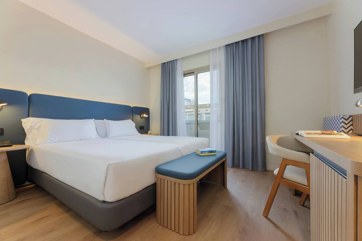 A hotel room with a bed, bedside table, bench, desk, chair, and a window with curtains.