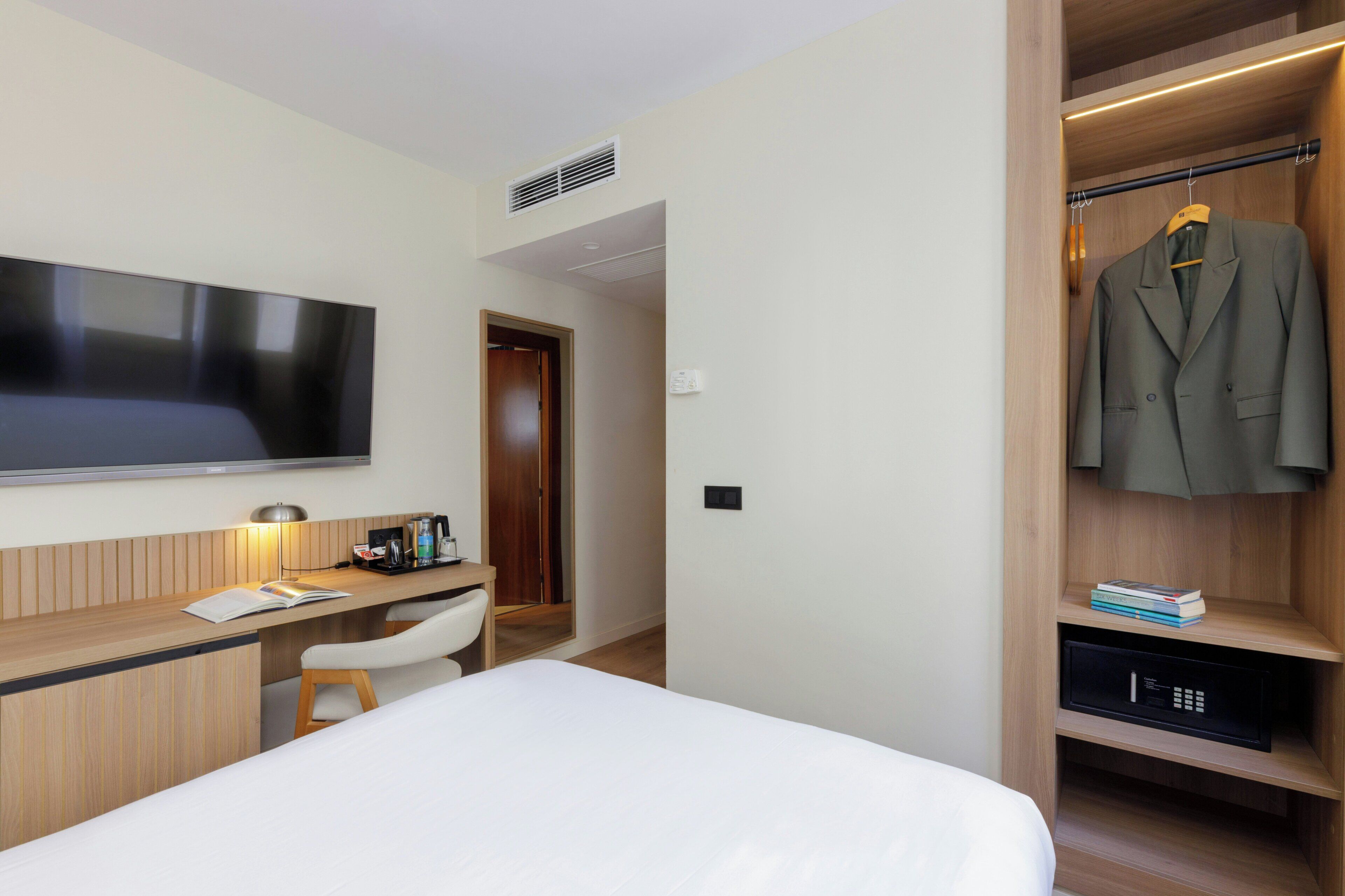 A modern hotel room with a flat-screen TV, wooden desk, white chair, and a walk-in closet with a hanging coat rack.