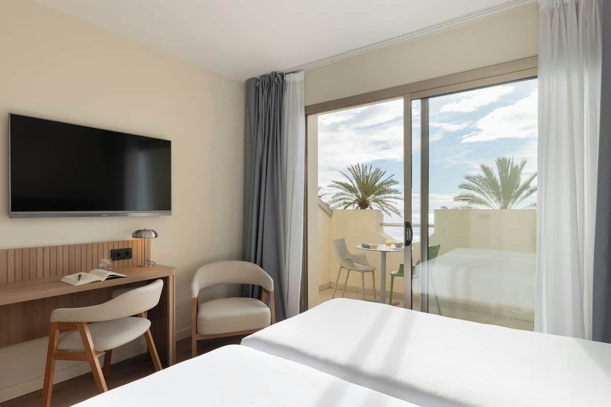 A hotel room with a bed, a desk with a chair, a TV, and a balcony with a table and chairs.