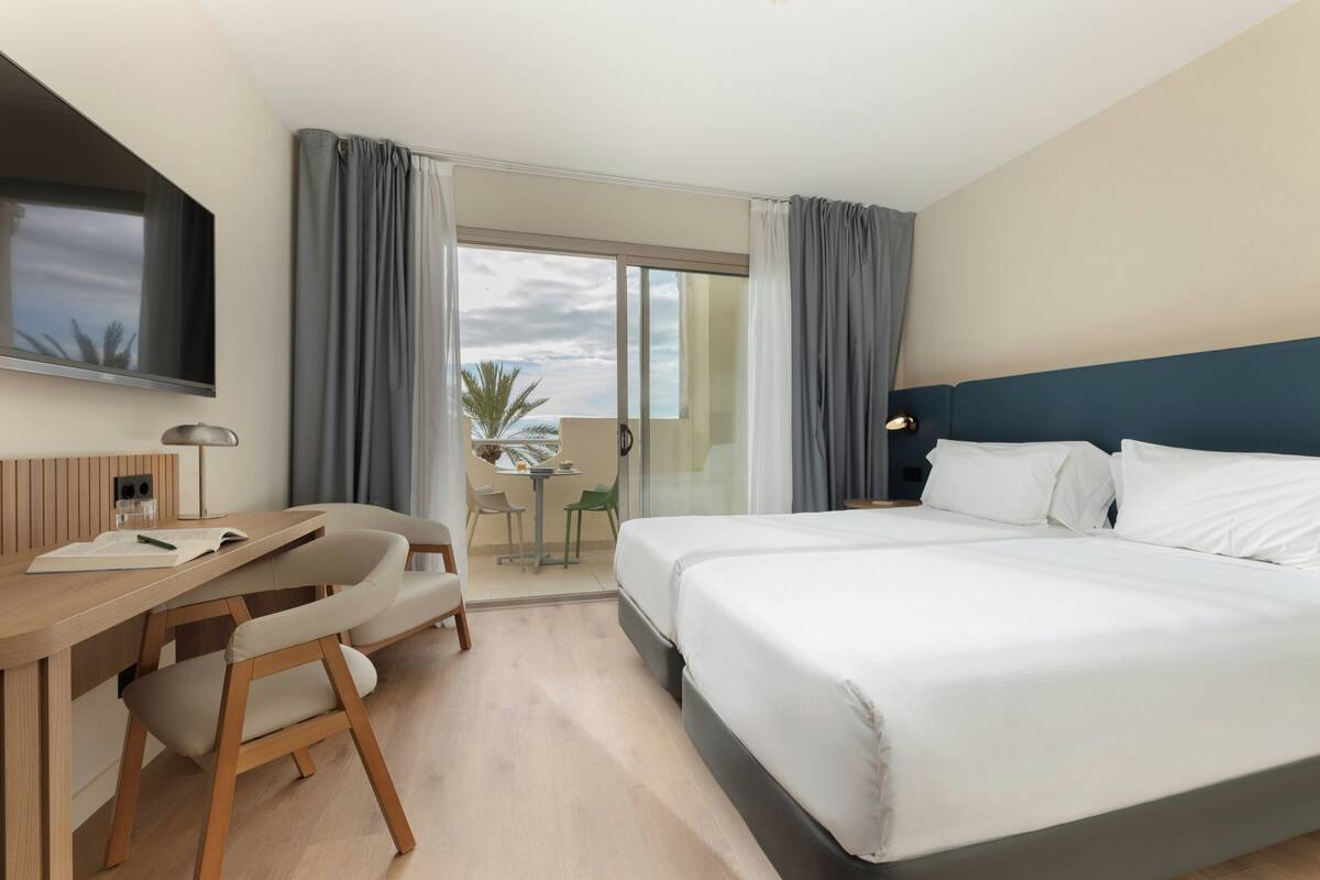 A hotel room with a bed, a desk, and a TV. There is a balcony with a table and chairs.