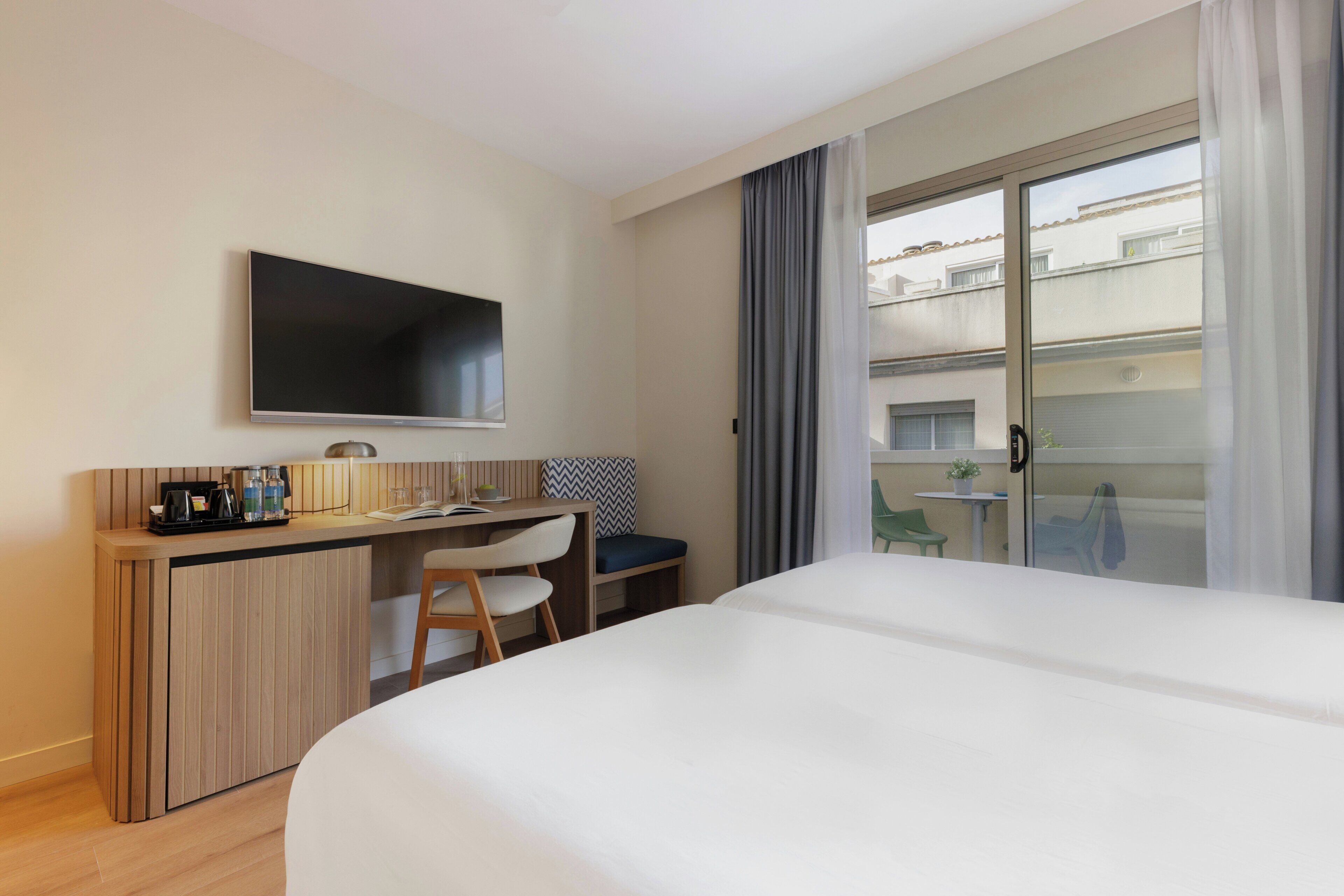 A hotel room with a bed, a desk with a chair, a TV, and a balcony with a view of buildings.