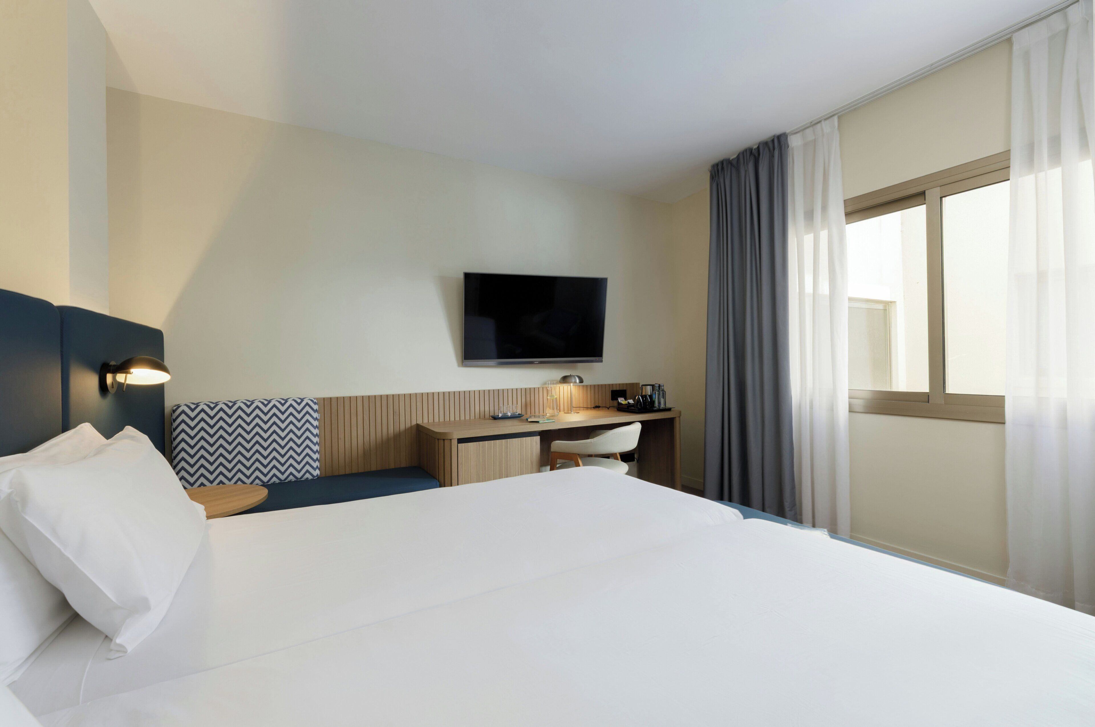 Una habitación de hotel con cama, escritorio, televisor y ventana con cortinas.