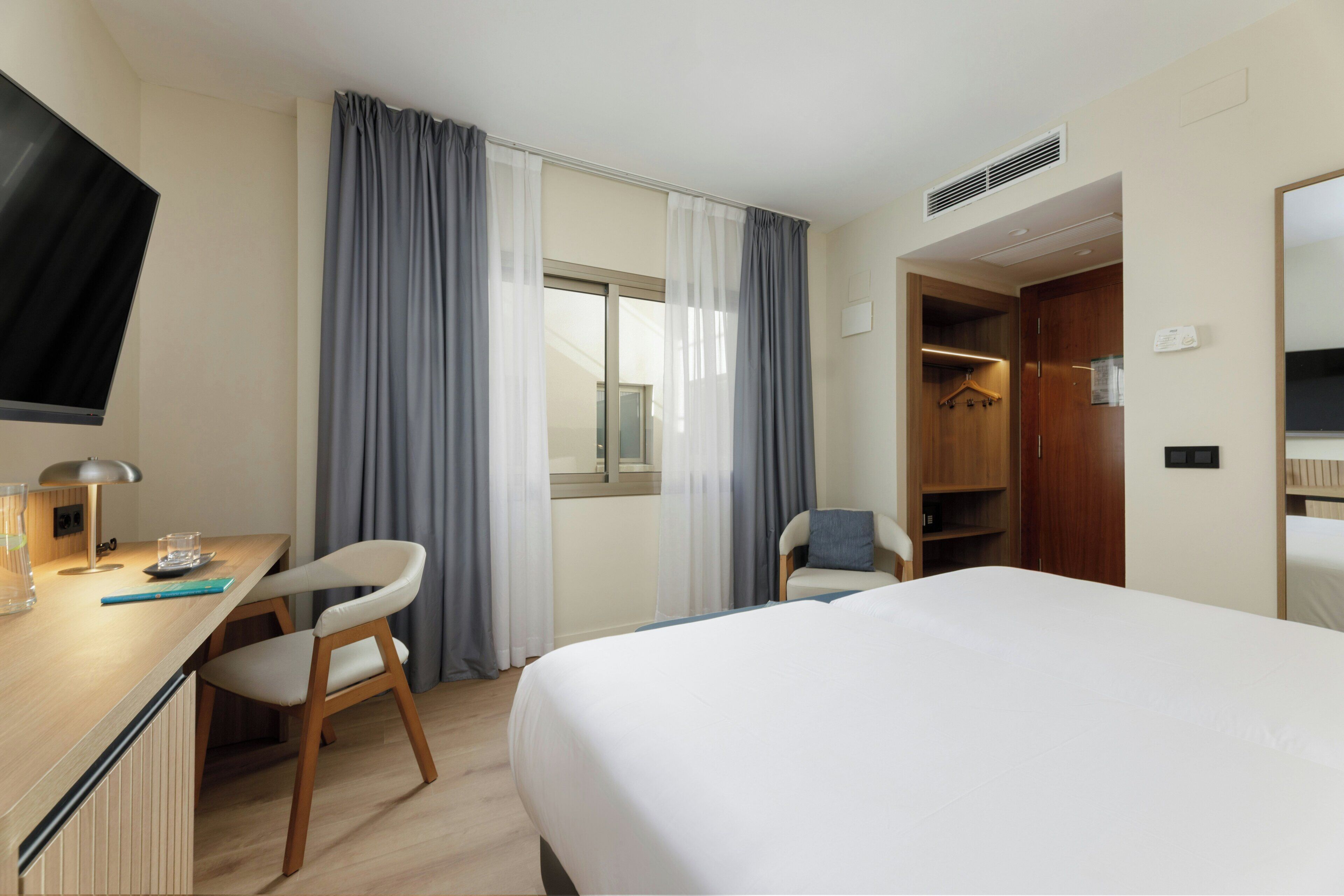 A hotel room with a bed, a desk with a chair, a TV, and a wardrobe.