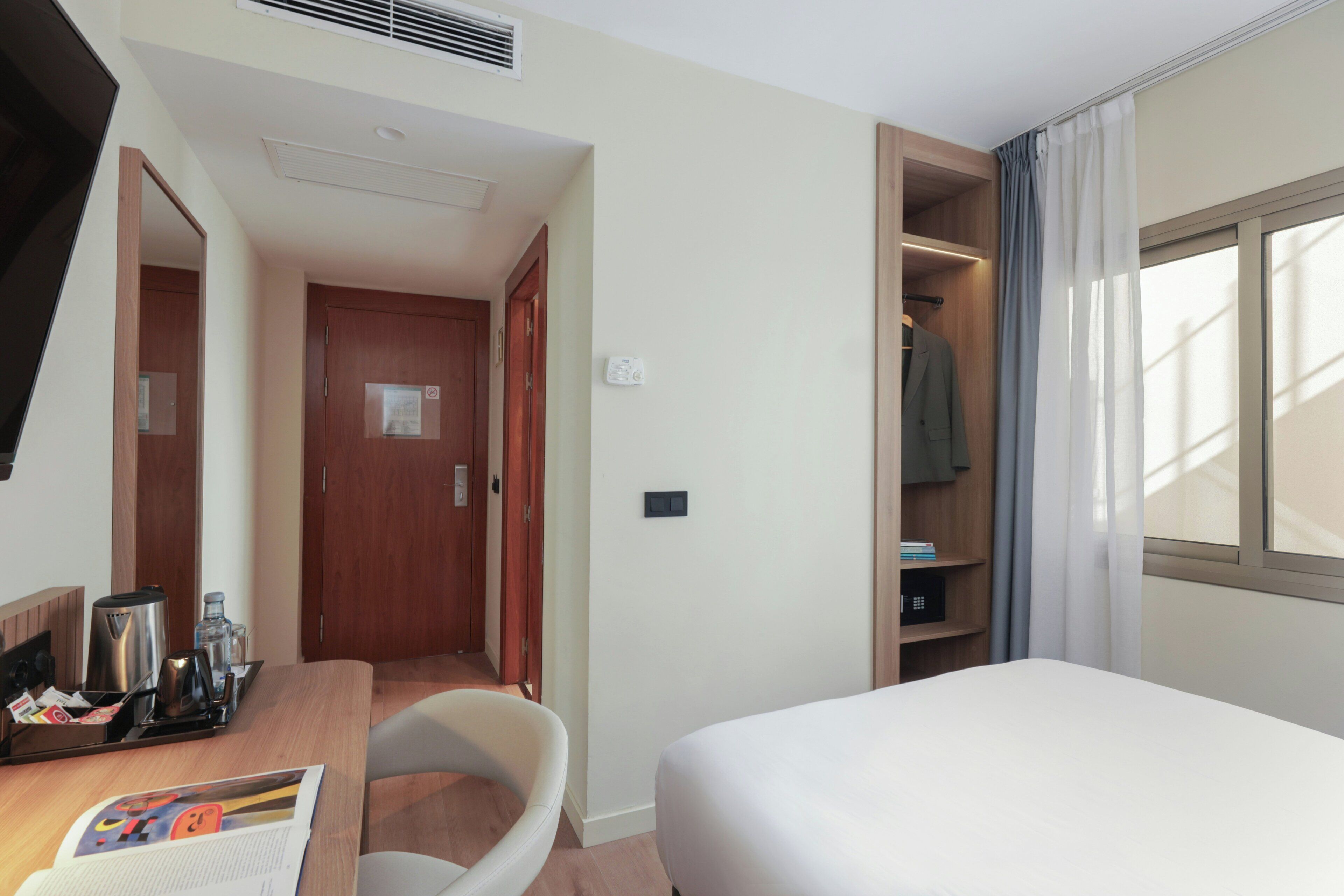 A hotel room with a bed, a desk with a chair, a wardrobe, and a window with curtains.