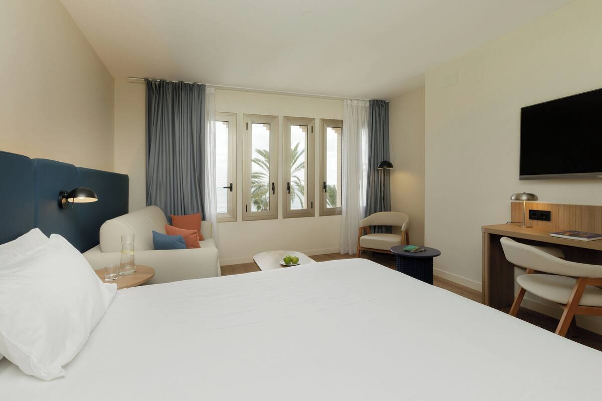 A hotel room with a large bed, a desk, a chair, a TV, and a window with curtains.