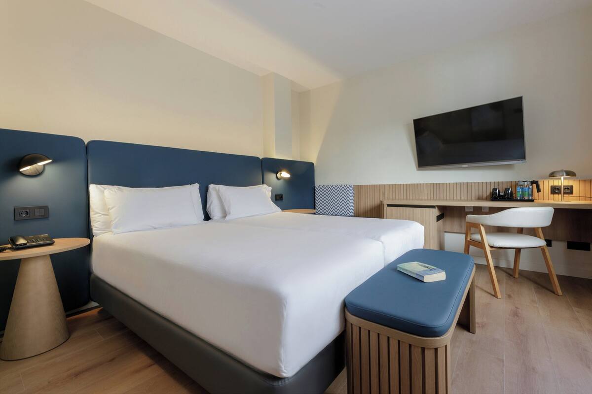 A modern hotel room with a large bed, bedside tables, a desk with a chair, a flat-screen TV, and a small reading area.