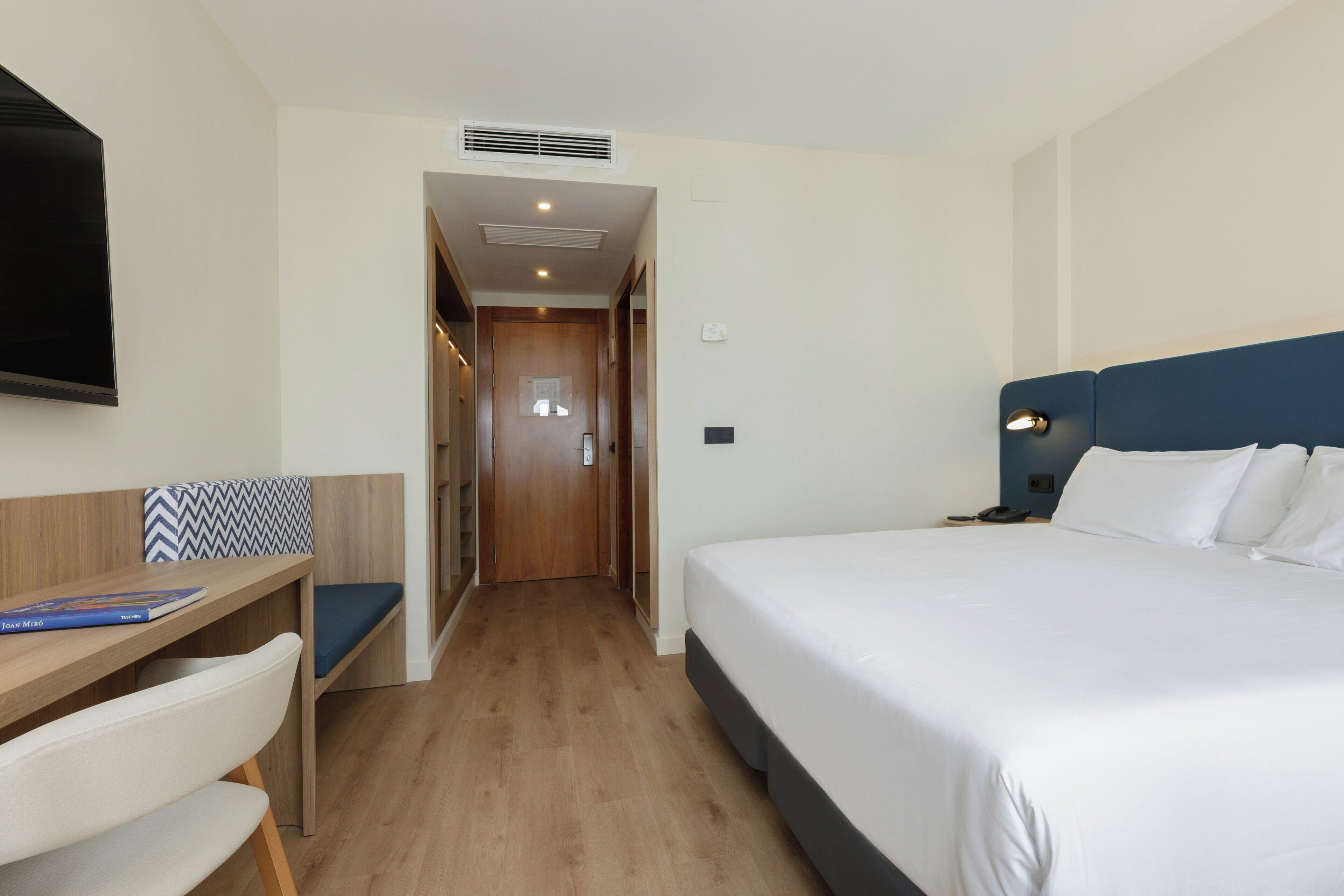 A hotel room with a bed, a desk with a chair, a television, and a door leading to a hallway.