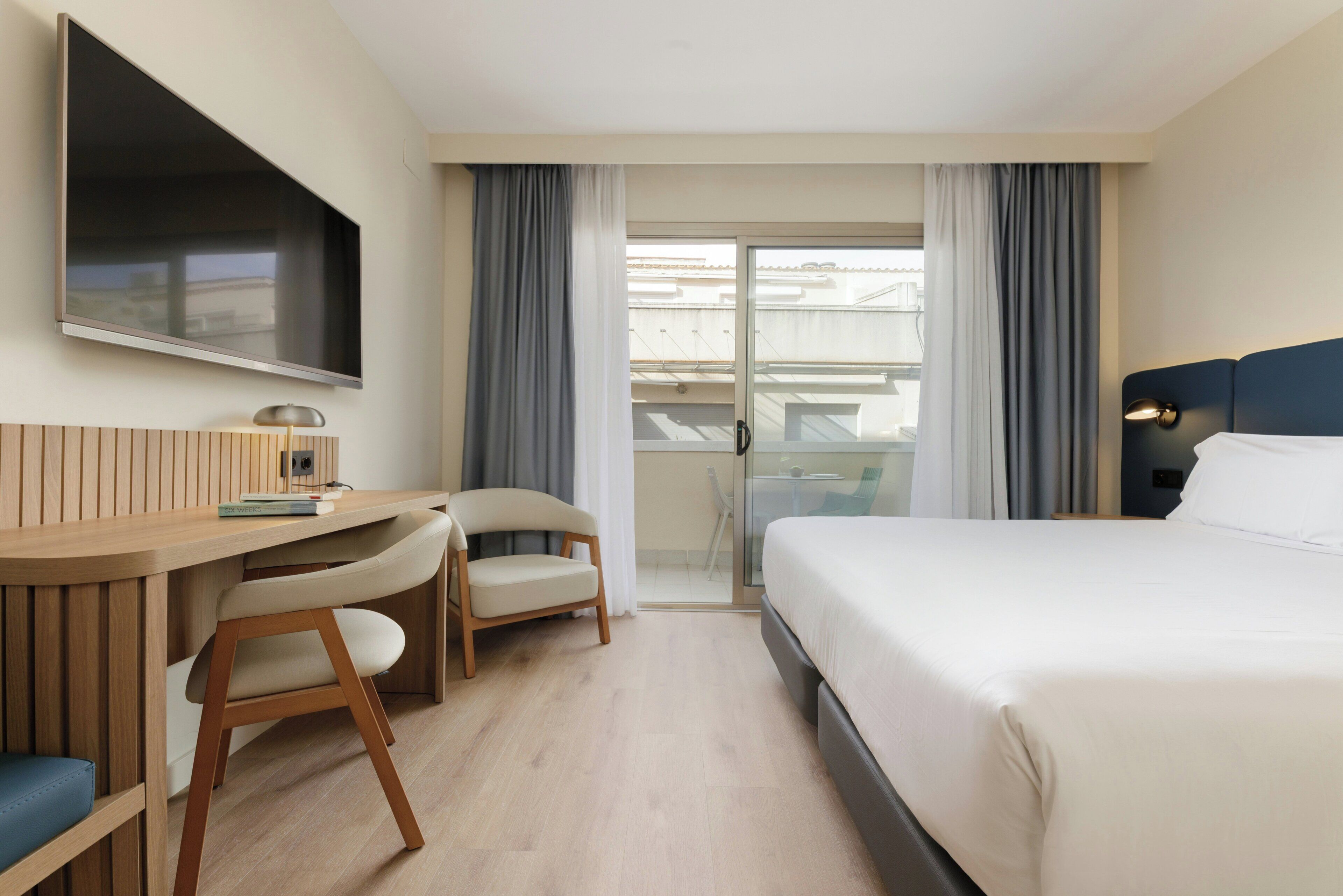 Una camera d'albergo con un letto, una scrivania con una sedia, una TV a parete e un balcone con tende.