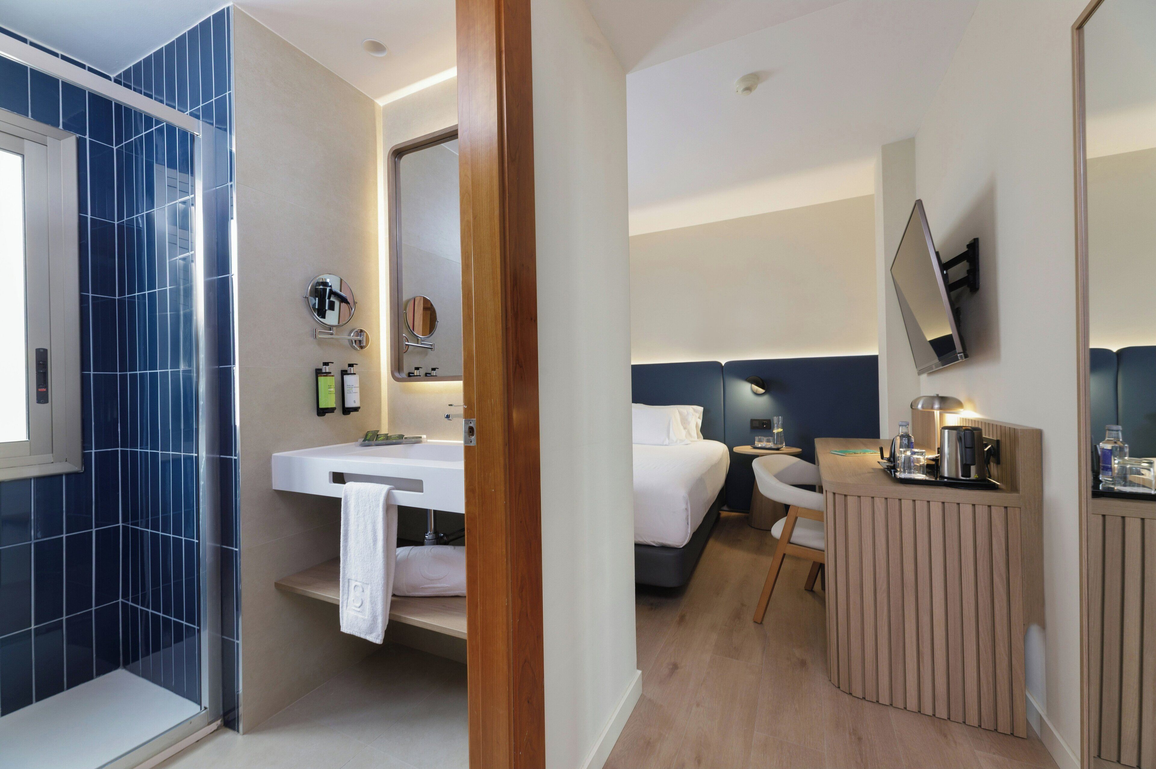 Una habitación de hotel moderna con ducha, lavamanos, cama, mesita de noche y televisor montado en la pared.
