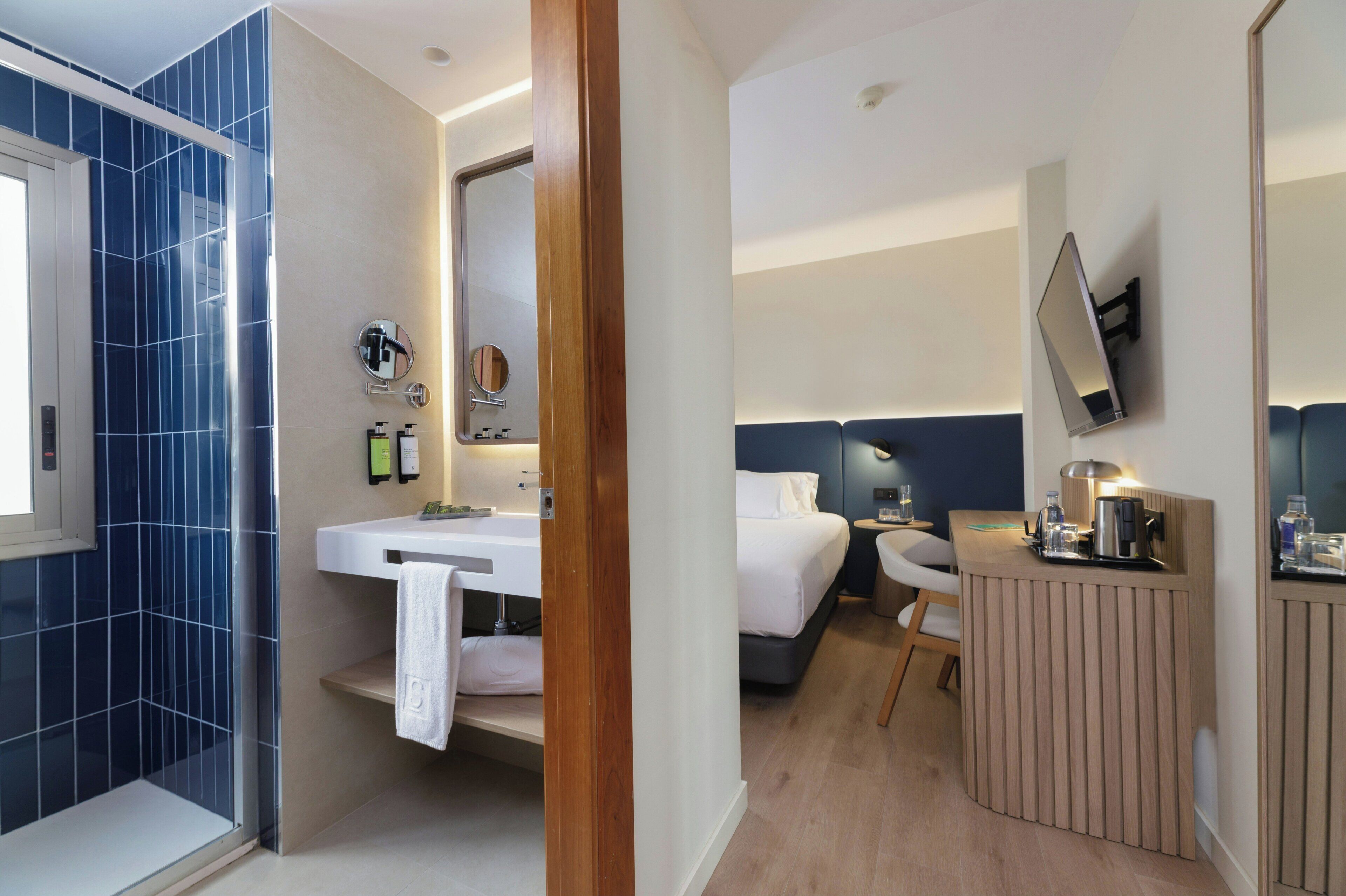 A modern hotel room with a shower, a sink, a bed, a bedside table, and a TV mounted on the wall.