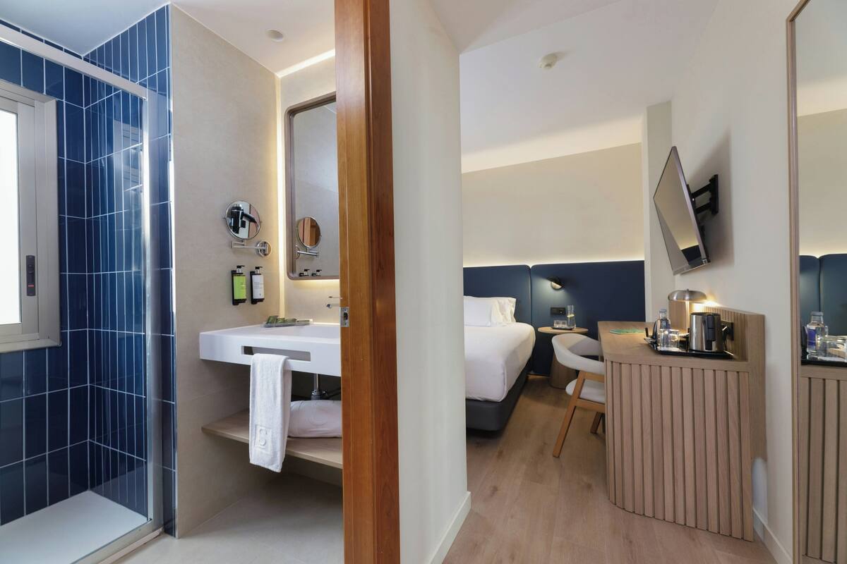 A modern hotel room with a shower, a sink, a bed, a bedside table, and a TV mounted on the wall.