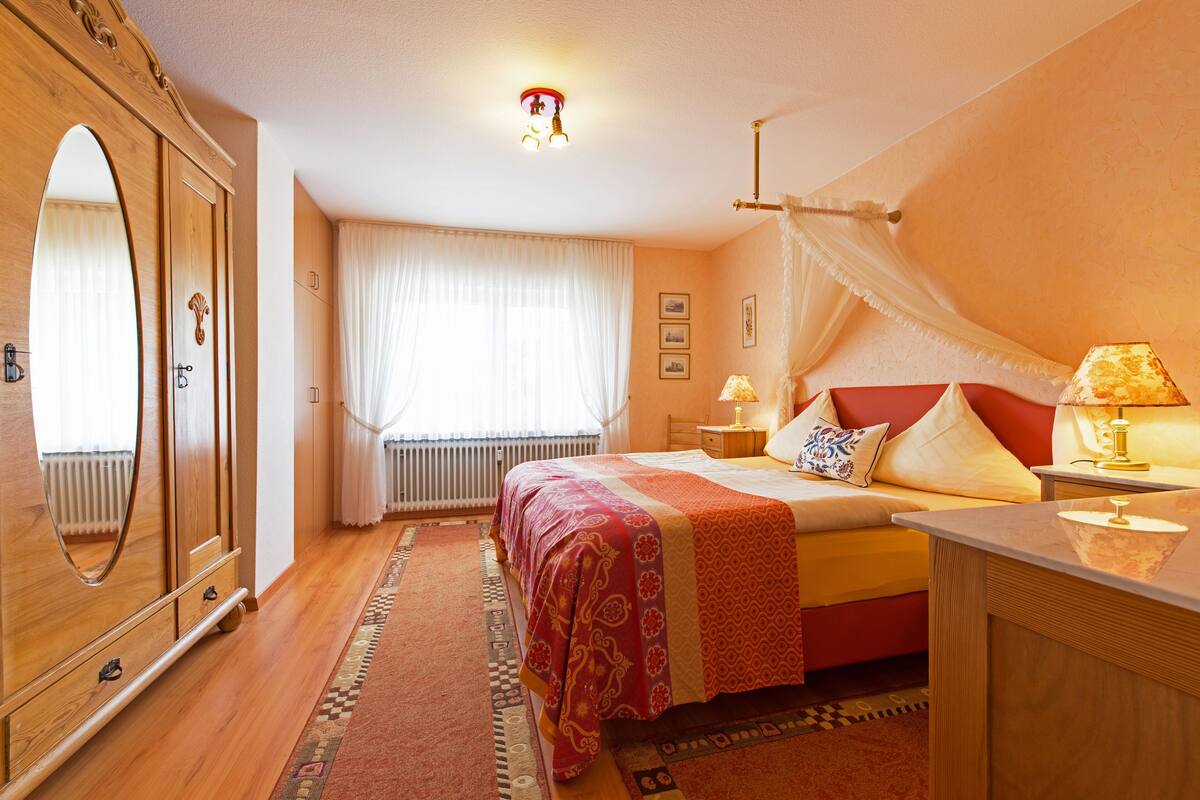 Une chambre avec un grand miroir, un lit recouvert d’une couverture rouge et blanche, deux lampes de chevet et une armoire en bois.