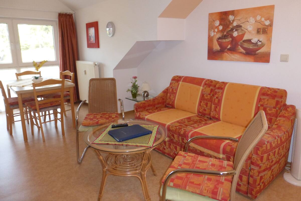 Un salon comprenant un canapé, des fauteuils, une table basse en verre, une table à manger avec des chaises, un tableau accroché au mur et un radiateur.