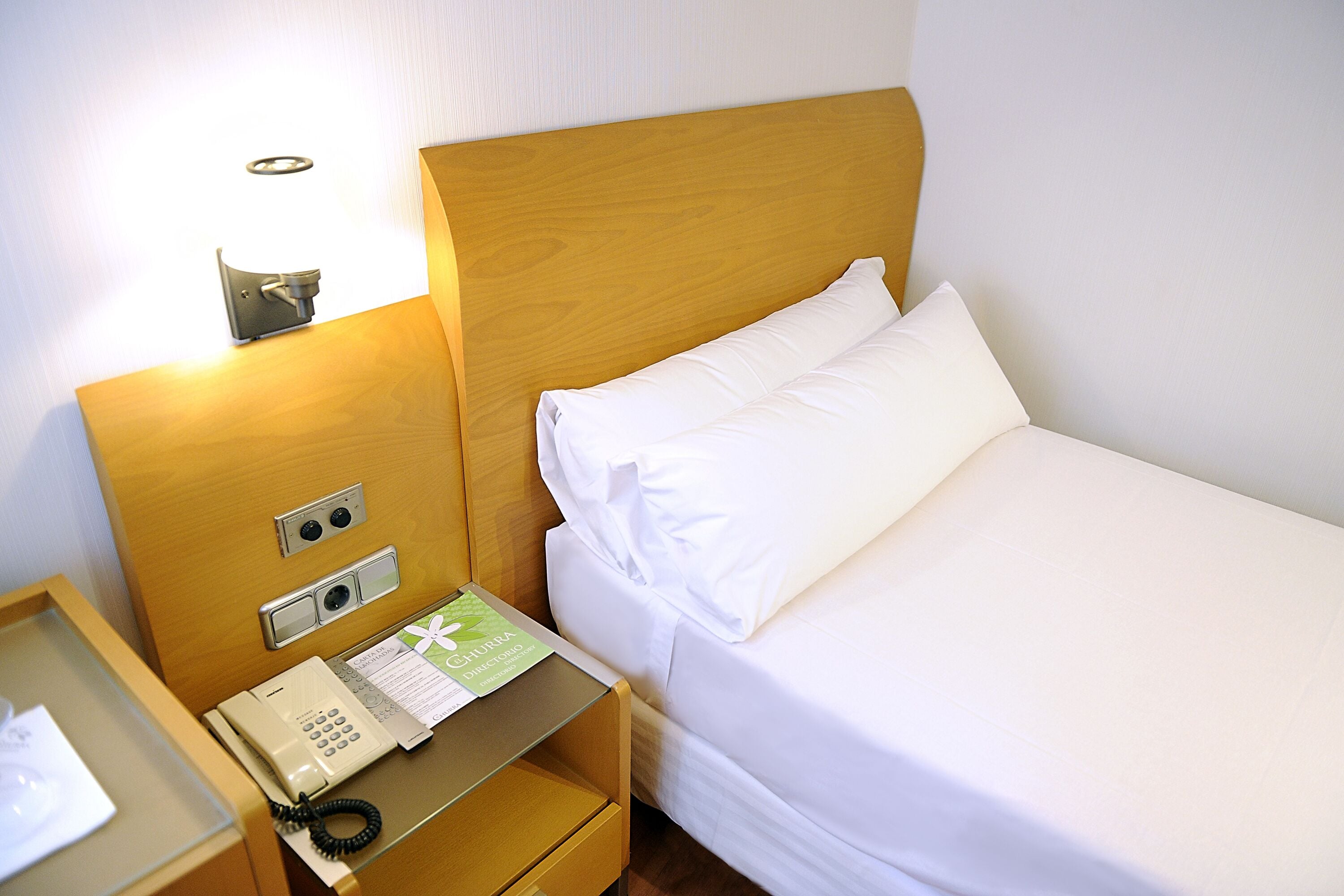 Una habitación de hotel con cabecera de madera, una mesita de noche con teléfono y una cama bien tendida.