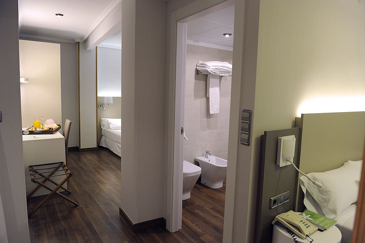 A hotel room with a wooden floor, a bed, a bathroom with a toilet and bidet, a small table with a tray, and a drying rack.