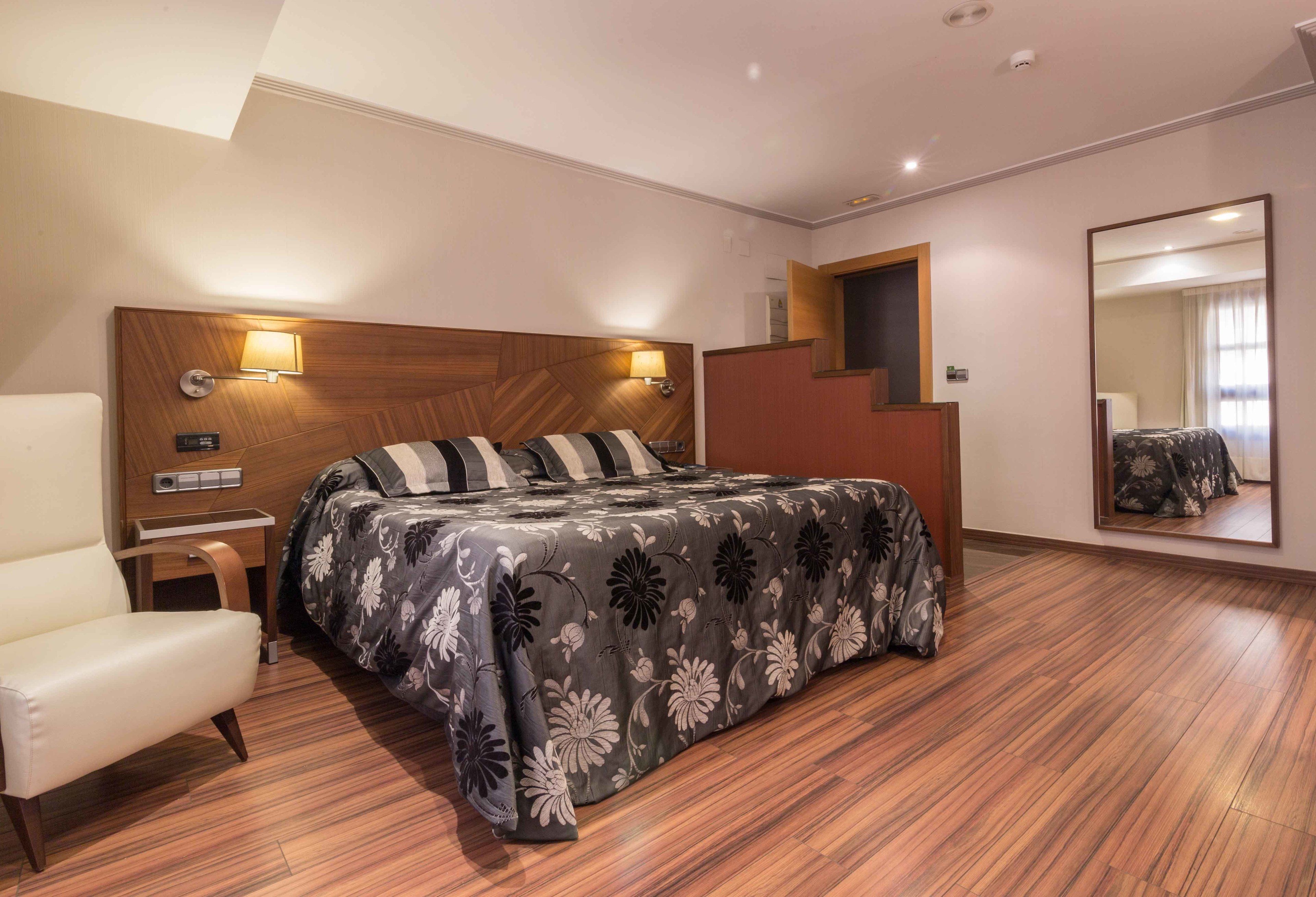 Una habitación de hotel moderna con cabecera de madera, cama con colcha floral, un sillón blanco, una mesita de noche y un espejo en la pared.