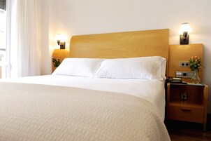 Una cama bien hecha con sábanas blancas, cabecera de madera, lámparas de noche, mesita de noche con un florero y teléfono de pared.