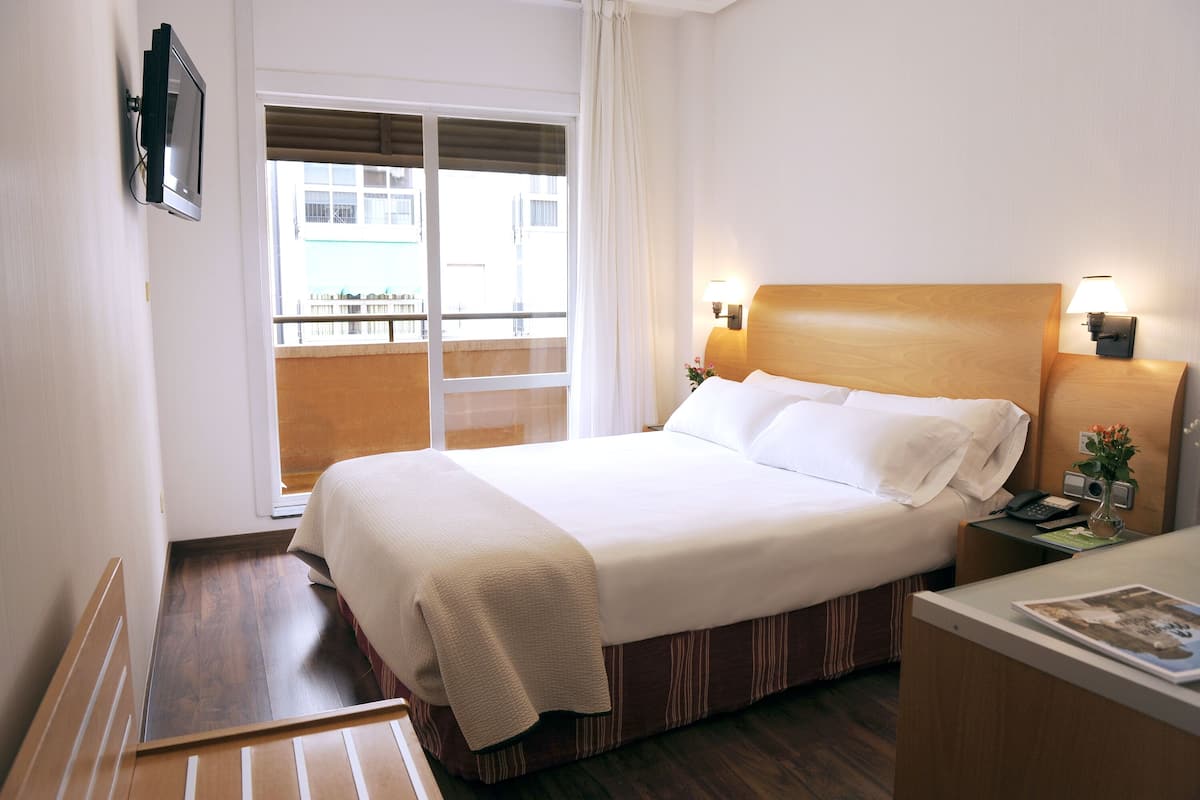 A hotel room with a bed, bedside table, lamp, and a view of the outside balcony.