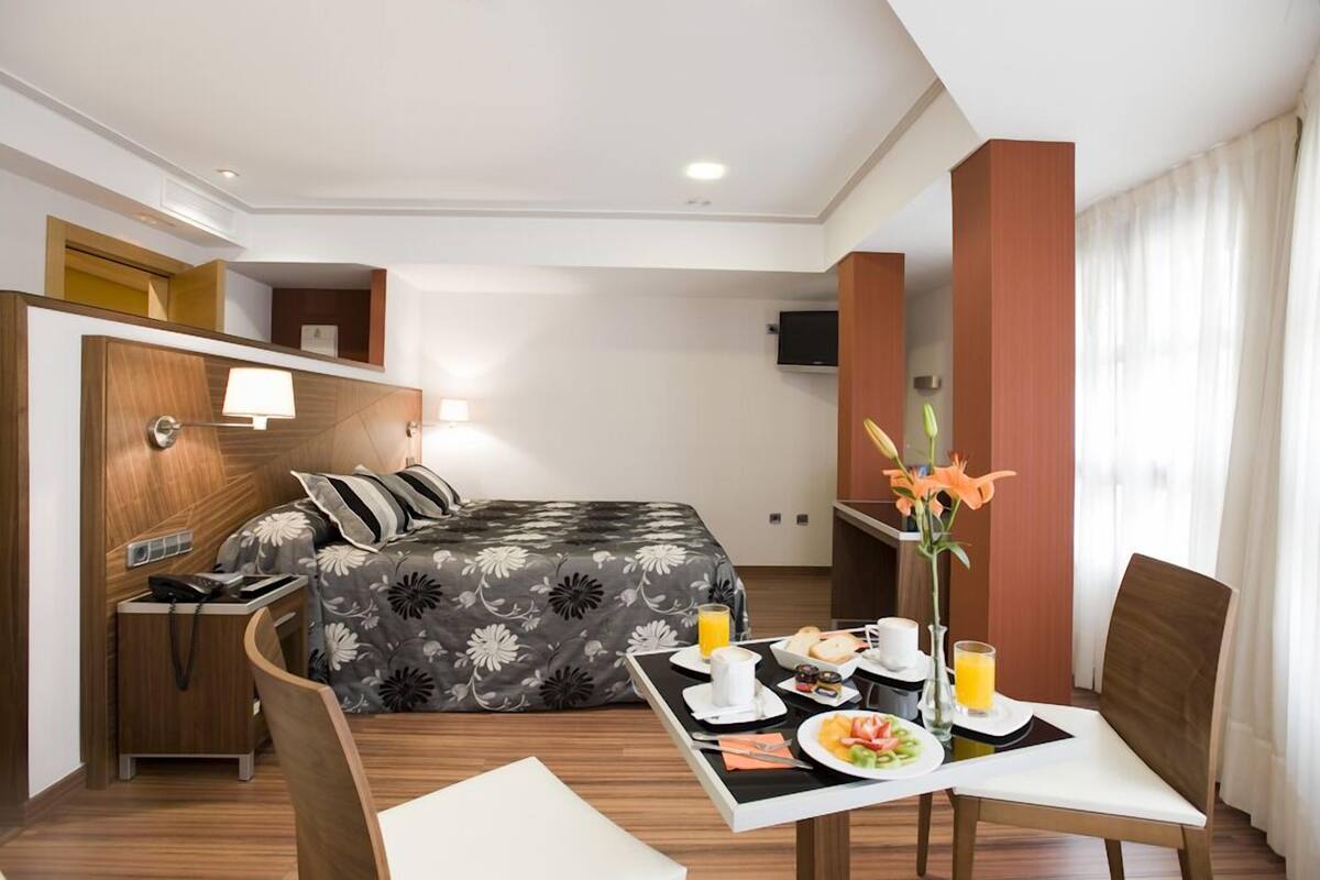 A hotel room with a bed, a desk with a lamp, a dining table with breakfast, and a TV on the wall.