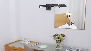 A compact hotel room with a wooden desk, a chair, a small vase with flowers, a mirror, and a wall-mounted lamp.