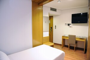 A hotel room with a bed, a desk, a chair, and a mounted TV.