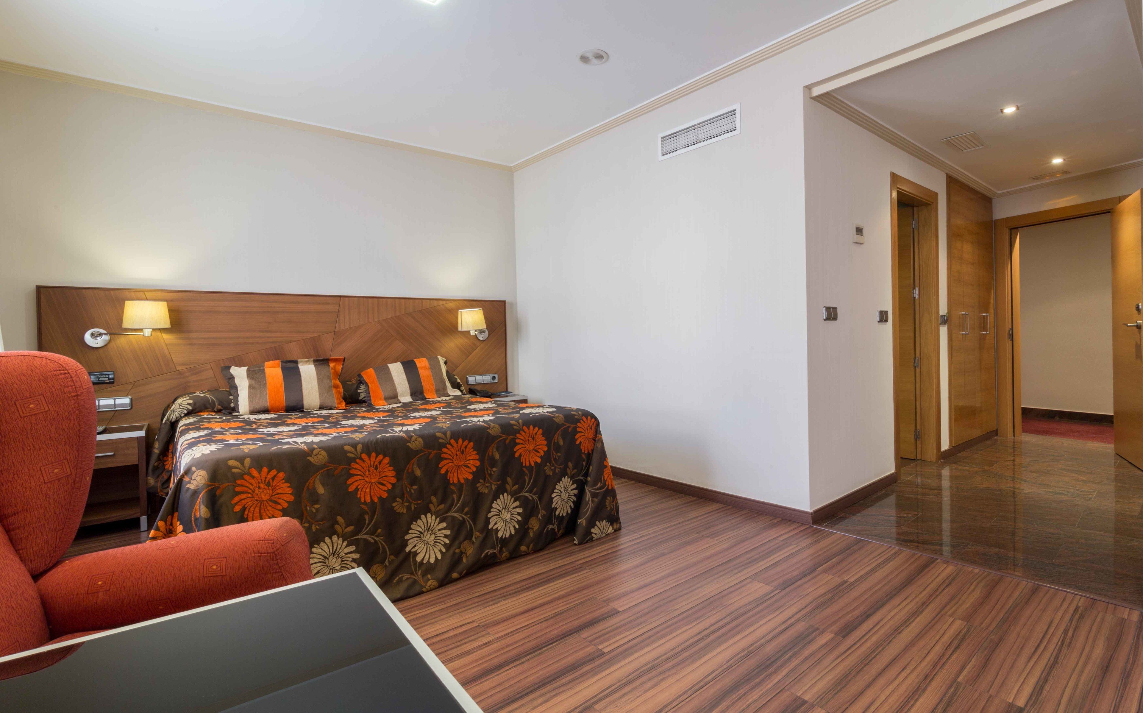 Une chambre d’hôtel avec une tête de lit en bois, un lit recouvert d’une literie à motifs floraux, un fauteuil rouge et un bureau avec une lampe.
