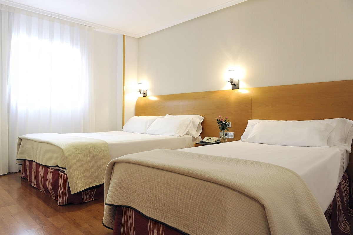 A hotel room with two beds, each with a wooden headboard, white bedding, and a bedside table with a lamp.