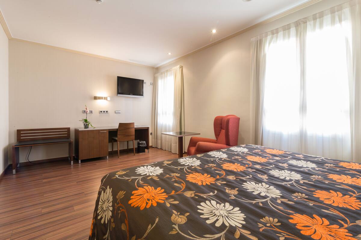 A hotel room with a bed, a desk, a chair, a TV, and a window with curtains.